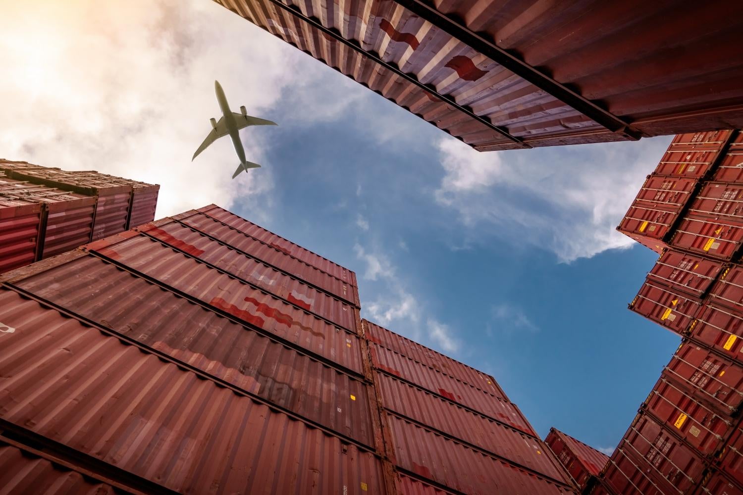 Skyward view of a plane through stacks of shipping containers