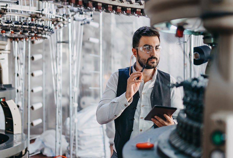 A man with dark hair and a beard, wearing goggles, stands in front of factory equipment.