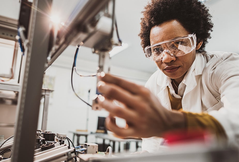 A Black woman wearing goggles and a lab coat touches a piece of equipment.