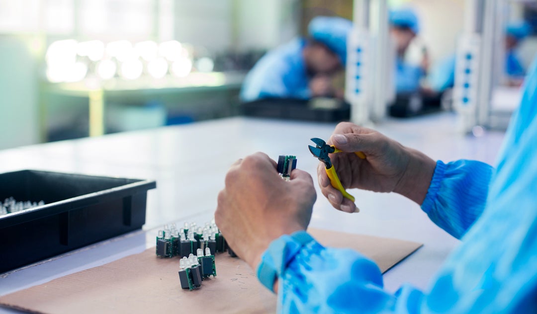 A closeup image of a person holding pliers and adjusting a semiconductors. In the background, other workers are adjusting semiconductors.