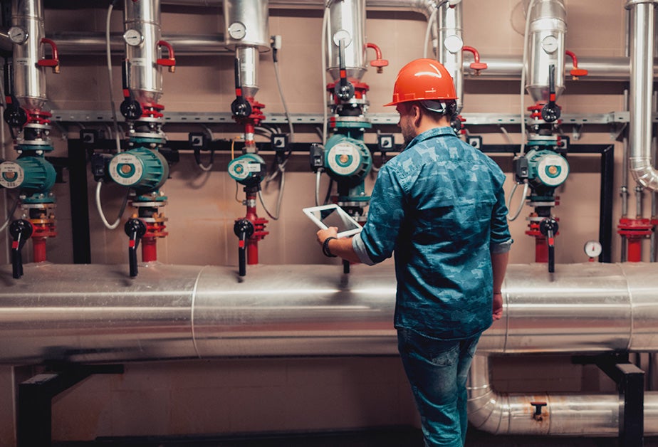 A man with an orange hard hat on his head and wearing a blue shirt and pants stands in front of a series of gauges.