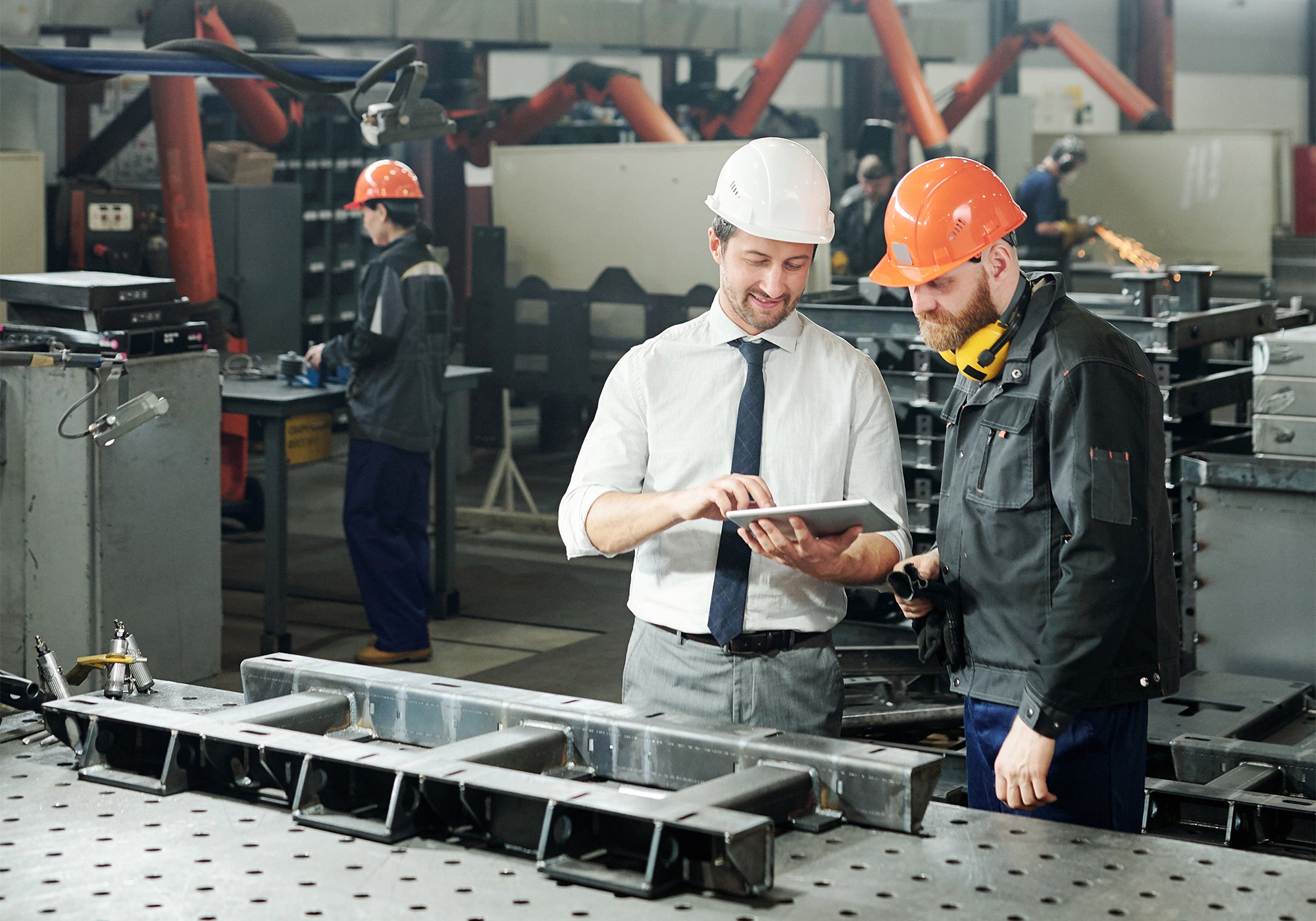 Two men in hard hats stand next to each other in a factory. On the table in front of them, there is a piece of metal equipment. In the background, there is another man with a hard hat.