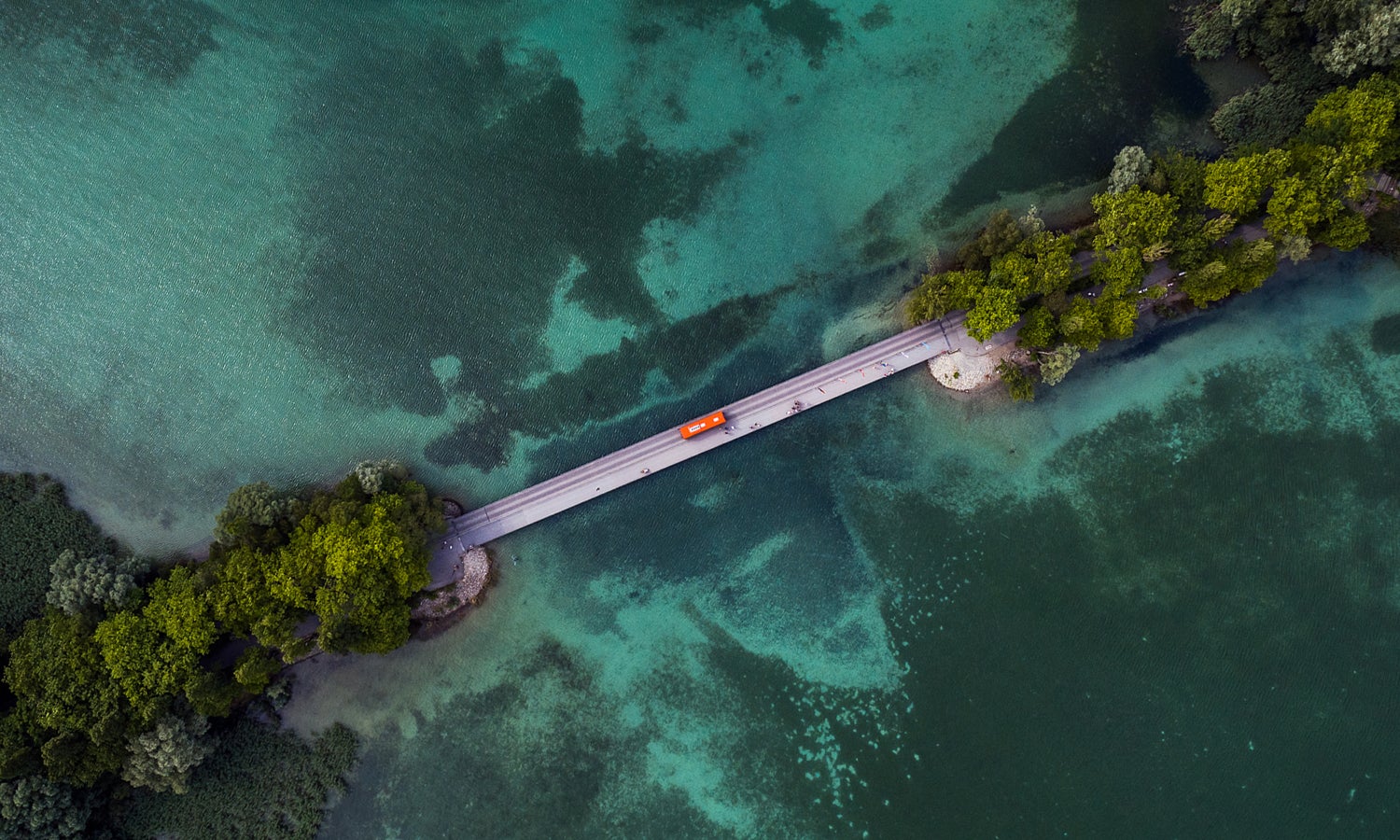 This is an aerial view of a narrow bridge connecting two thin pieces of forest and a road.