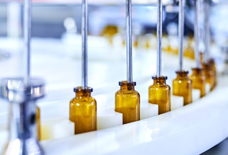 On an assembly line, brown glass bottles are filled by tubes.