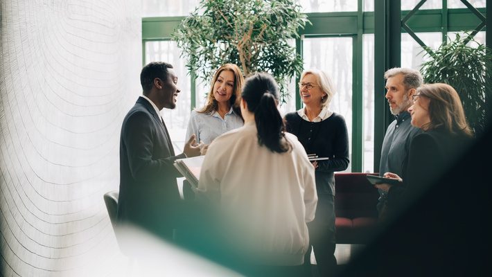 A diverse group of professionals smiling and conversing in a bright, modern office space, symbolizing inclusion, collaboration, and community impact.