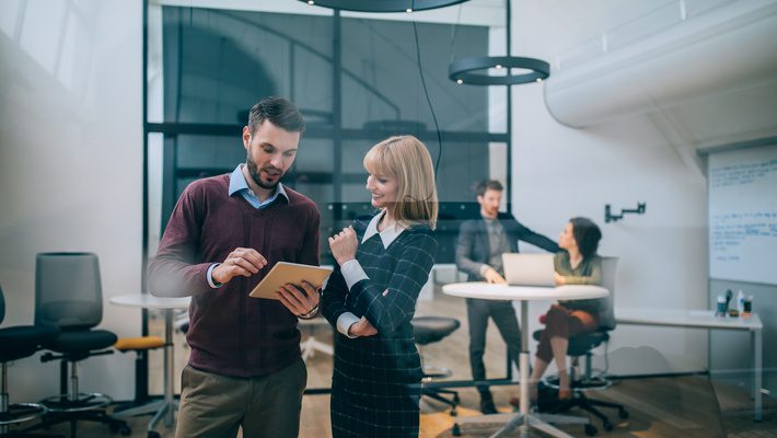 Business professionals discussing compliance strategy on a digital tablet in a modern office setting, with team collaboration in the background.